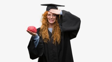 Giyen genç redhead öğrenci kadın mezun Tekdüzen holding beyin kafasında, utanç ve sürpriz yüz ile şok el ile vurguladı öfkeli ve sinirli. Korku ve hata için üzgün.