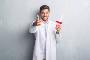 Handsome young doctor man over grey grunge wall holding diploma happy with big smile doing ok sign, thumb up with fingers, excellent sign clipart