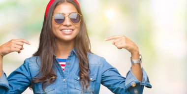 Genç güzel Arap kadın yüzünde gülümseme ile kendinden emin görünüyorsun, kendini gururlu ve mutlu parmakları ile işaret izole arka plan üzerinde güneş gözlüğü takıyor.