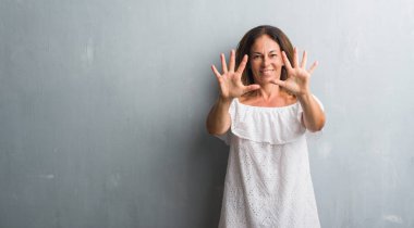 Gösterilen ve parmakları ile işaret gri grunge duvarın üzerinden duran orta yaş İspanyol kadın kendinden emin ve mutlu gülümseyerek süre on numara.