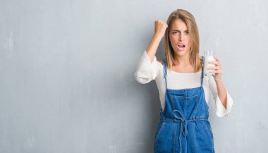 Güzel genç kadın grunge gri duvar driking bir bardak süt rahatsız ve boşa çıkarmak ile öfke, çılgın ve yükseltilmiş eliyle diye bağıran öfke kavramı bağırarak.