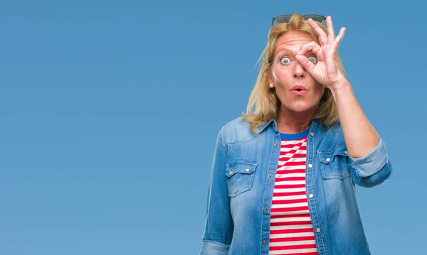 Middle age blonde woman over isolated background doing ok gesture shocked with surprised face, eye looking through fingers. Unbelieving expression.