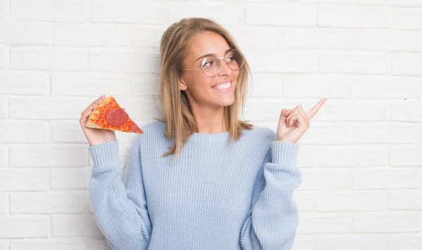 白いレンガの壁側の手と指で指す非常に幸せなピザのスライスを食べる以上の美しい若い女性 — ストック写真