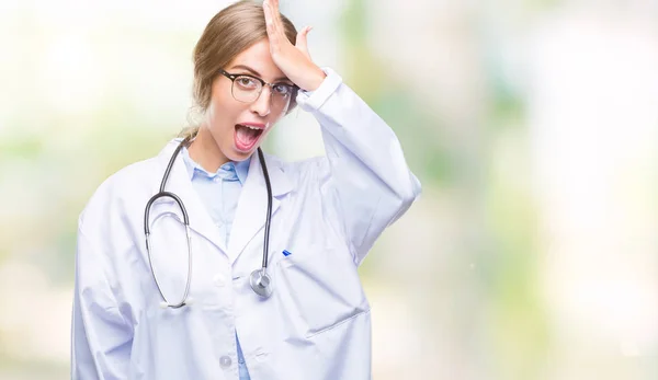 Beautiful young blonde doctor woman wearing medical uniform over isolated background surprised with hand on head for mistake, remember error. Forgot, bad memory concept.