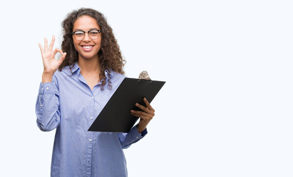 Young hispanic business woman holding clipboard doing ok sign with fingers, excellent symbol