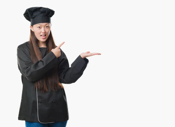 驚くし 手とを提示 指で指している間カメラに笑顔のシェフの制服を着て孤立の背景の上の若い中国の女性 — ストック写真