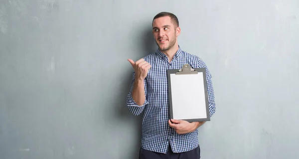 Young Caucasian Man Grey Grunge Wall Holding Clipboard Pointing Showing — Stock Photo, Image