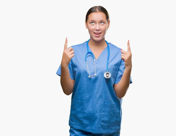 若い白人医師女性隔離された背景に医療制服を着てびっくり驚いて見上げると指で指していると腕を上げた — ストック写真