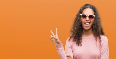 Güzel genç İspanyol kadın mutlu yüz zafer işareti yaparken kameraya göz kırpıyor ile gülümseyen güneş gözlüğü takıyor. İki numaralı.