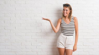 Beyaz tuğla duvar neşeli sunulması ve kameraya bakarak avucunun ile işaret gülümseyen genç güzel sarışın kadın.