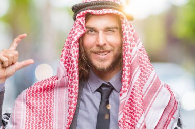 Genç yakışıklı Arap adam uzun saçlı izole arka plan gülümseyen ve süre seyir ve kamera boyutu işaret parmaklarıyla yapıyor el ile kendinden emin eller üzerinde keffiyeh giyiyor. Ölçü kavramı.
