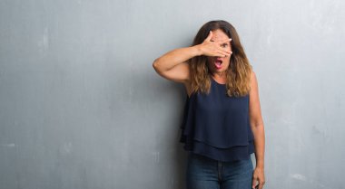 Mahcup ifadeyle parmaklarımın arasından seyir gri grunge duvar yüz ve gözler el ile kapsayan şok bakmak üzerinde duran orta yaş İspanyol kadın.