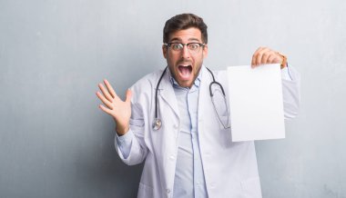 Yakışıklı genç doktor adam gri grunge duvar büyük gülümseme ile çığlık zaferini kutluyor ve ellerini kaldırdı sayfa boş sözleşme çok mutlu ve heyecanlı, kazanan ifade holding