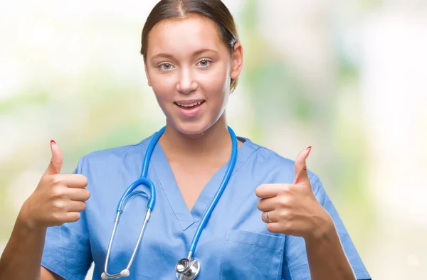 年轻的白种人医生妇女穿医制服在孤立的背景成功标志做积极的手势与手 竖起大拇指微笑和快乐 用欢快的表情看着相机 胜利者的手势 — 图库照片