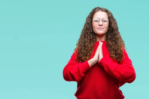Beautiful brunette curly hair young girl wearing glasses and winter sweater over isolated background praying with hands together asking for forgiveness smiling confident.