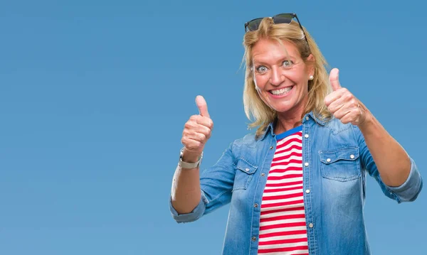 Middle age blonde woman over isolated background approving doing positive gesture with hand, thumbs up smiling and happy for success. Looking at the camera, winner gesture.