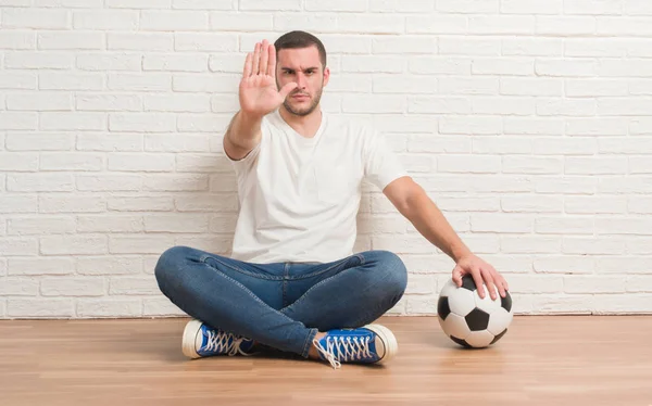 年轻的白种人坐在白色砖墙举行足球足球与张开手做停止标志以严肃和自信的表达 防御姿态 — 图库照片