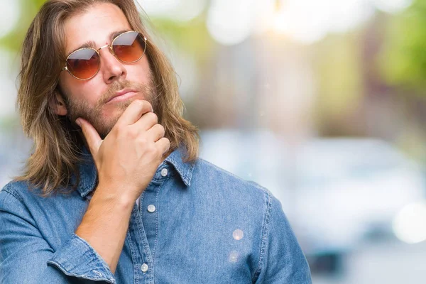 Young handsome man with long hair wearing sunglasses over isolated background with hand on chin thinking about question, pensive expression. Smiling with thoughtful face. Doubt concept.