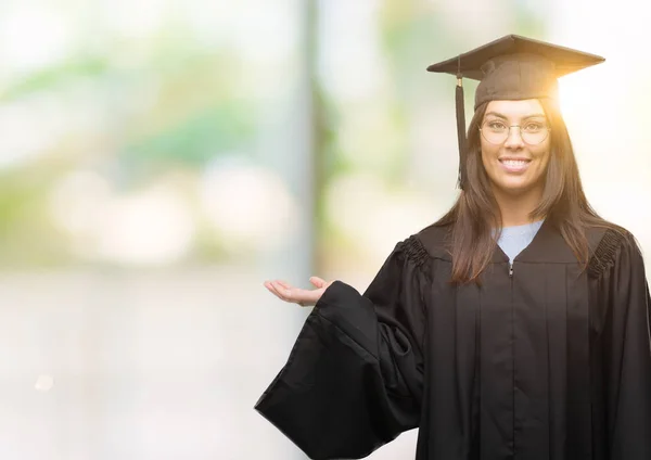 年轻的拉美裔妇女穿着毕业帽和均匀微笑欢快地呈现和指向与手掌看着相机 — 图库照片