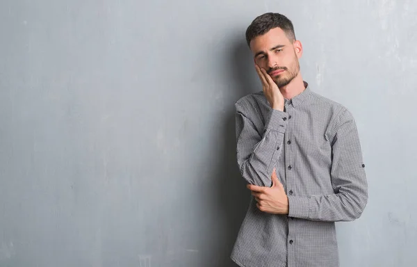 年轻的男子站在灰色的垃圾墙思考看起来累了 厌倦了与交叉武器抑郁症问题 — 图库照片