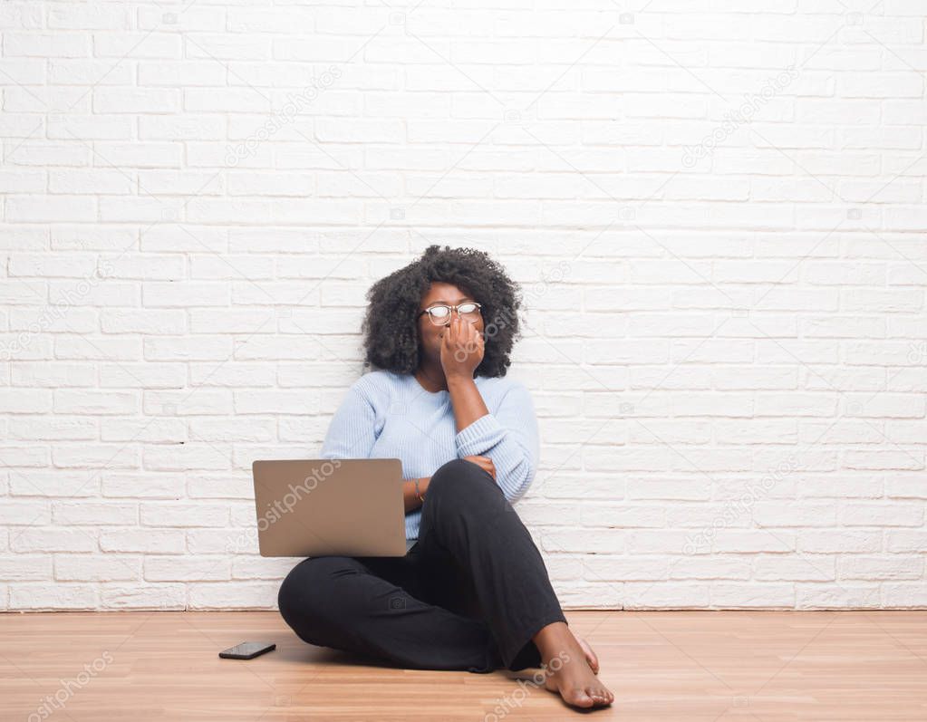 Young african american woman sitting on the floor using laptop at home looking stressed and nervous with hands on mouth biting nails. Anxiety problem.