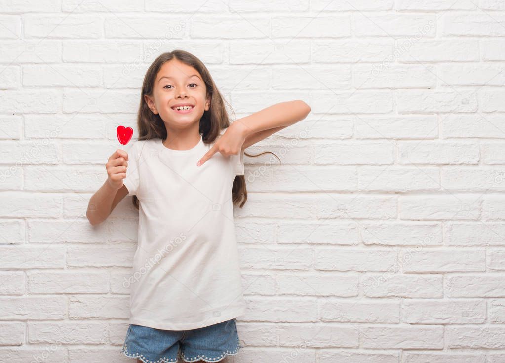 Young hispanic kid over white brick wall eating red heart lollipop candy with surprise face pointing finger to himself