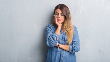 Grunge gri duvar düşünme seyir yorgun ve çapraz kolları olan depresyon sorunları ile sıkılmış gözlüklü Genç Yetişkin kadın.