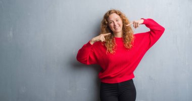 Emin gösterilen ve işaret parmakları diş ve ağız ile gülümseyen genç redhead gri grunge duvar giyen üzerinde kırmızı kadın kazak. Sağlık kavramı.