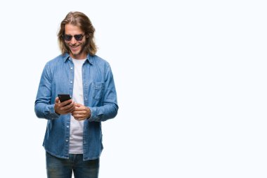 İzole arka plan ayakta ve diş gösterilen kendine güvenen bir gülümsemeyle gülümseyerek mutlu bir yüz ile smartphone kullanarak ileti gönderme üzerinde uzun saçlı yakışıklı delikanlı