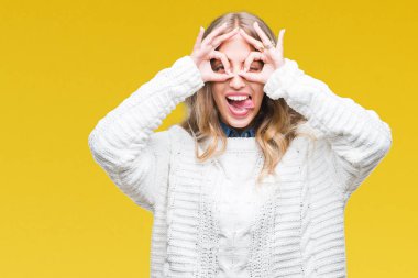 Dilini, parmaklarının arasından baktığını yapışmasını dürbün gibi ok jest yapıyor izole arka plan üzerinde kış kazak giyen güzel genç sarışın kadın. Deli ifade.