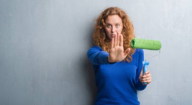 Gri grunge duvar boya silindiri açık eliyle dur işareti ile ciddi ve kendine güvenen ifade, savunma jest yapıyor tutan genç Kızıl saçlı kadın