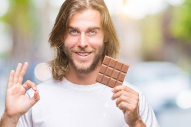 Uzun saç beslenme ile yakışıklı delikanlı çikolata bar ok işareti parmaklar, mükemmel sembolü ile yapıyor izole arka plan üzerinde