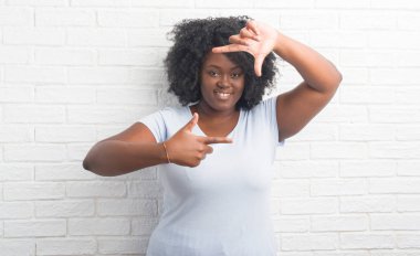 Genç Afro-Amerikan artı boyutu kadın elle yapma çerçeve ve parmakları ile mutlu yüz gülümseyen beyaz tuğla duvarın üzerinden. Yaratıcılık ve Fotoğrafçılık kavramı.