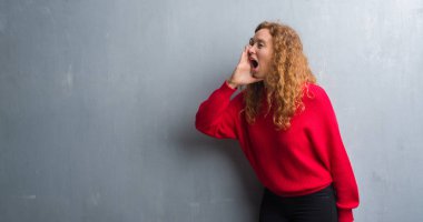 Kırmızı kazaklı bağırarak ve ağzı üzerinde el ile yan için çığlık gri grunge duvarın üzerinden genç Kızıl saçlı kadın. İletişim kavramı.