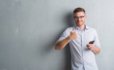 Genç Kızıl saçlı adam gri grunge duvar gönderen mesaj ok işareti, başparmak ile parmak, mükemmel işareti yapıyor kocaman bir gülümseme ile smartphone mutlu kullanarak