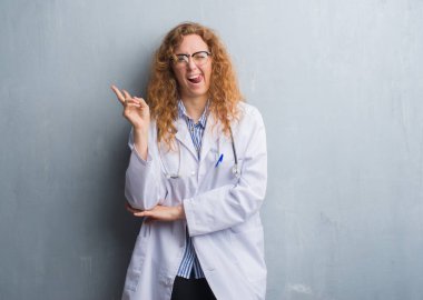Genç redhead doktor kadın mutlu yüz zafer işareti yaparken kameraya göz kırpıyor ile gülümseyen bir palto giyiyor gri grunge duvarın üzerinden. İki numaralı.