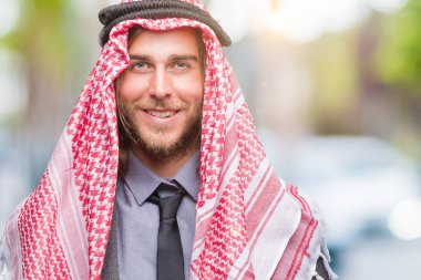 Genç yakışıklı Arap adam uzun saçlı yüzünde mutlu ve güzel bir gülümseme ile izole arka plan üzerinde keffiyeh giyiyor. Şanslı kişi.
