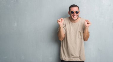 Zafer kutluyor gülümseyen Genç beyaz adam silah ile başarı için heyecanlı güneş gözlüğü takıyor gri grunge duvarın üzerinden kaldırdı. Kazanan kavramı.