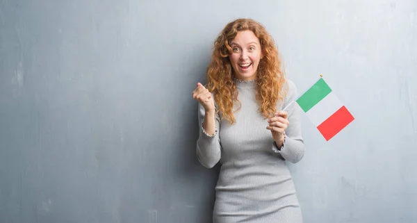 Joven Pelirroja Sobre Pared Grunge Gris Sosteniendo Bandera Italia Gritando —  Fotos de Stock