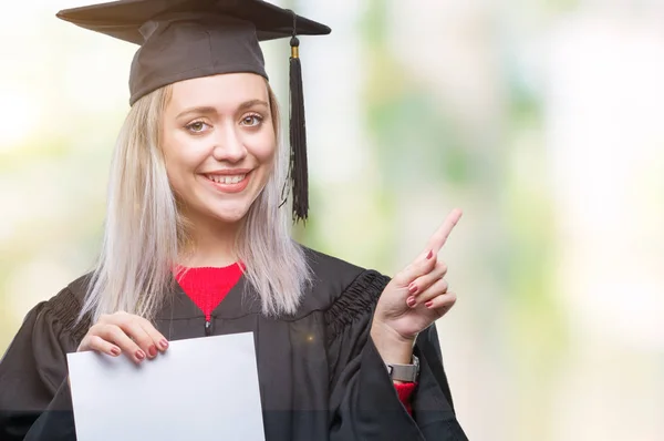 若いブロンドの女性側の手と指で指す非常に幸せな孤立の背景の上の学位を保持する大学院の制服を着て — ストック写真