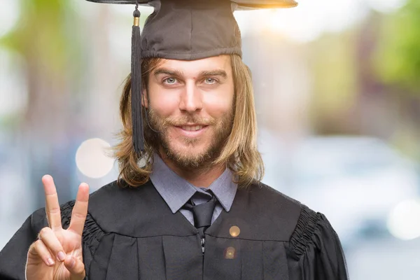 若いハンサムな勝利のサインをやって指を表示するカメラを笑みを浮かべて孤立の背景に長い髪を持つ男を卒業しました — ストック写真