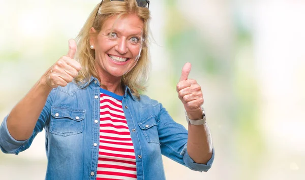 Middle age blonde woman over isolated background approving doing positive gesture with hand, thumbs up smiling and happy for success. Looking at the camera, winner gesture.