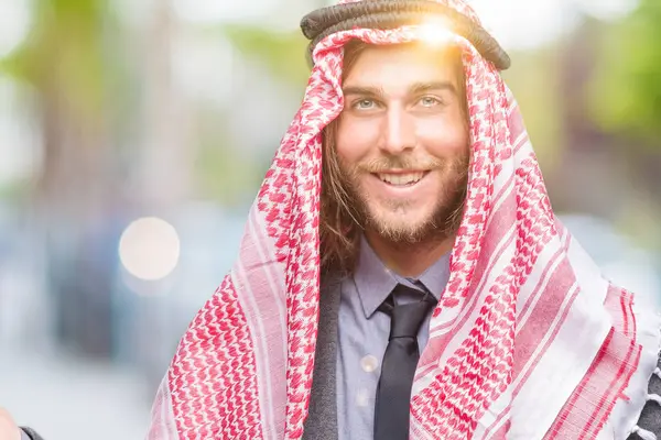 驚くし 手とを提示 指で指している間カメラに笑顔孤立の背景にクーフィーヤを着て長い髪の若いハンサムなアラビア人 — ストック写真
