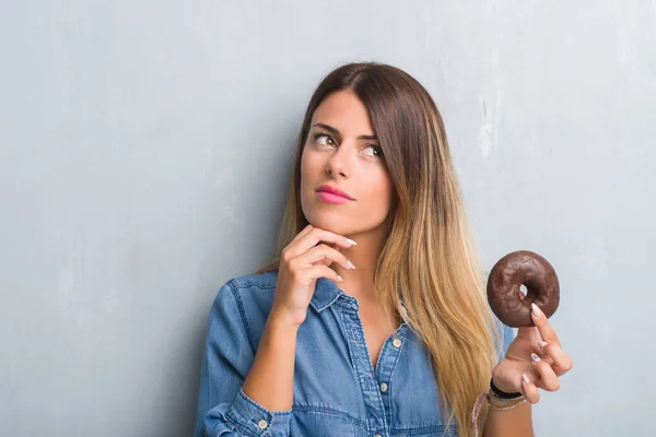 チョコレートを灰色のグランジの壁を食べる以上の若い成人女性ドーナツ深刻な顔を考えて質問 非常に混乱して考え — ストック写真