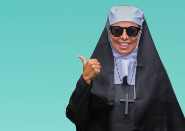 Orta yaş üst düzey Katolik Rahibe kadın arıyorum ve başparmak yanında yukarıyı mutlu yüz ile gülümseyen izole arka plan üzerinde güneş gözlüğü takıyor.