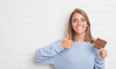 Güzel ve genç birbayan yeme çikolata mutlu ok işareti, başparmak ile parmak, mükemmel işareti yapıyor kocaman bir gülümseme ile beyaz tuğla duvarın üzerinden