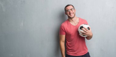 Genç beyaz adam ayakta ve diş gösterilen kendine güvenen bir gülümsemeyle gülümseyerek mutlu bir yüz ile futbol futbol topu tutan gri grunge duvarın üzerinden