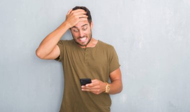 Yakışıklı genç adam gri grunge duvar manifatura elle kafasına, utanç ve sürpriz yüz ile şok vurguladı smartphone kullanarak bir ileti öfkeli ve sinirli. Korku ve hata için üzgün.