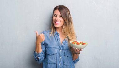 Genç Yetişkin kadın sağlıklı domates salatası ile mutlu büyük yemek gri grunge duvarın üzerinden ok işareti yaparken gülümse, parmaklarıyla, mükemmel işaret parmak