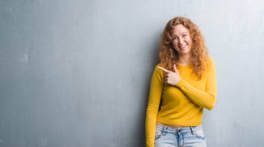 Gri grunge duvar neşeli bir gülümseme ile el ve parmak kadar kameraya bakarak yüzündeki mutlu ve doğal ifade ile yan ile işaret yüz genç Kızıl saçlı kadın.